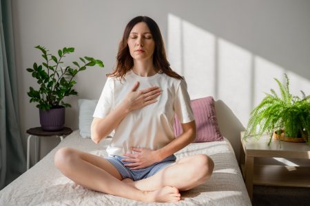 Pretty female sitting on bed in lotus posture with hands on her chest and belly doing pranayama techniques Doing yoga at home Self care and positive emotions
